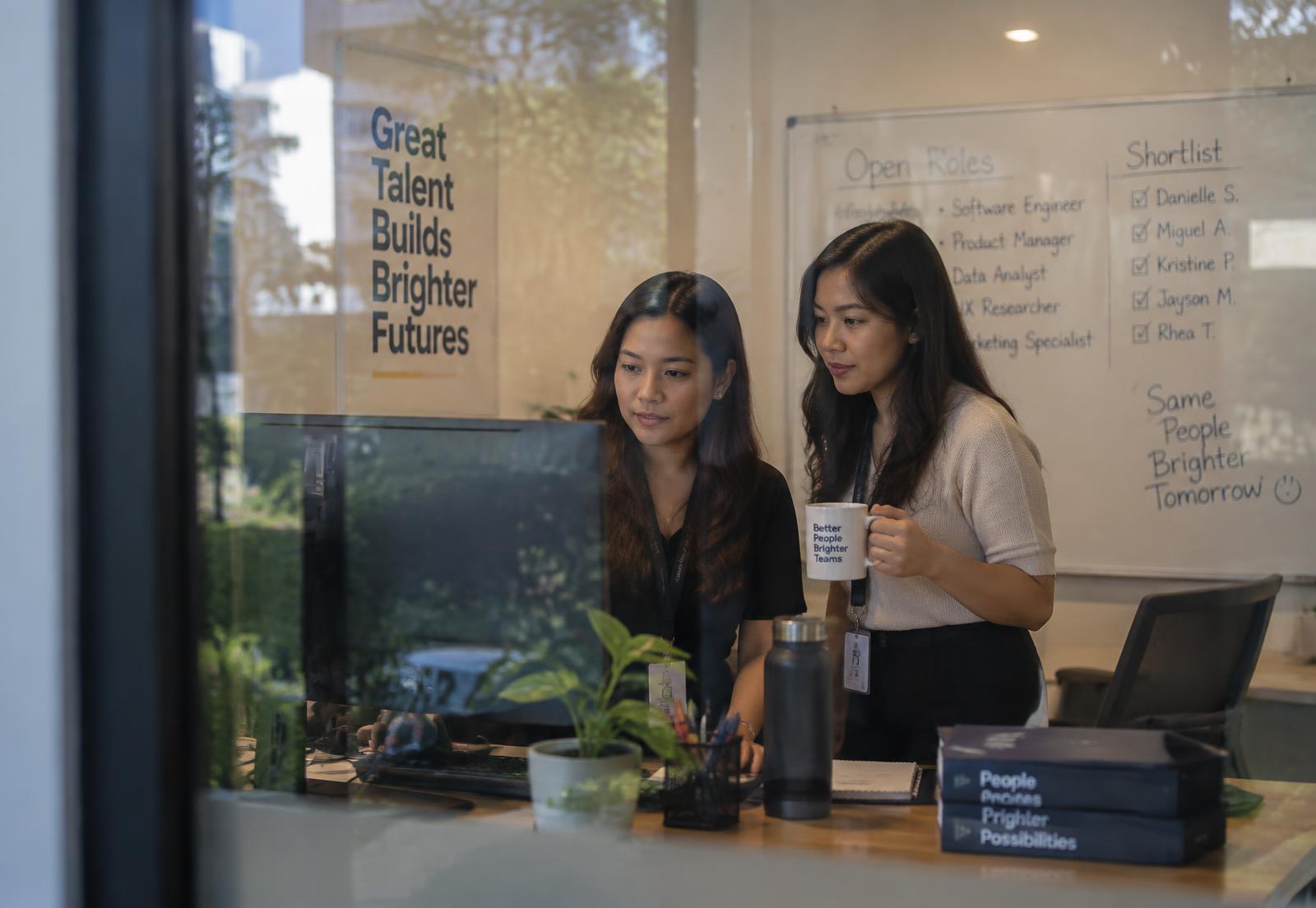 Two recruiters building a shortlist at a monitor beside an open roles whiteboard